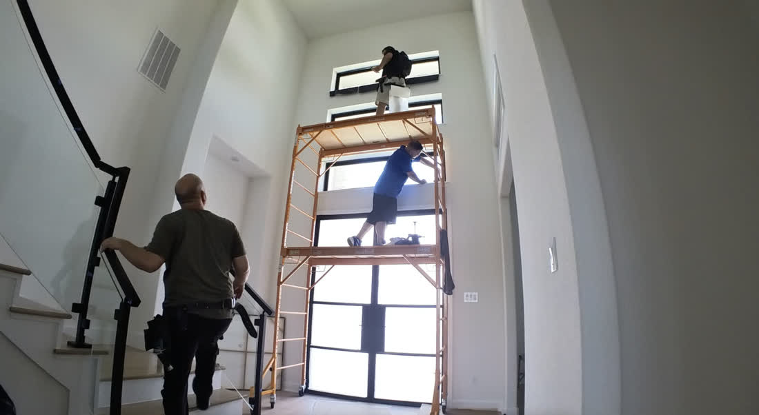 Two installers on a scaffold applying film to a two-story window above a staircase